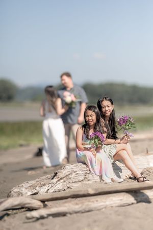 Group sitting on driftwood at beach holding purple flower bouquets on sunny day.