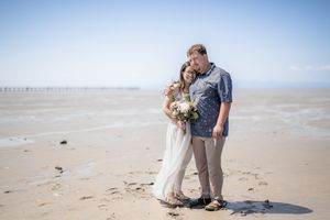 A couple shares a tender moment on a misty beach with a floral bouquet in hand while standing on the sandy shore.