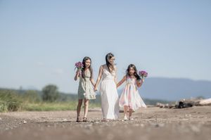 Three figures in light-colored dresses walk hand-in-hand along a dusty path with mountains in the background.