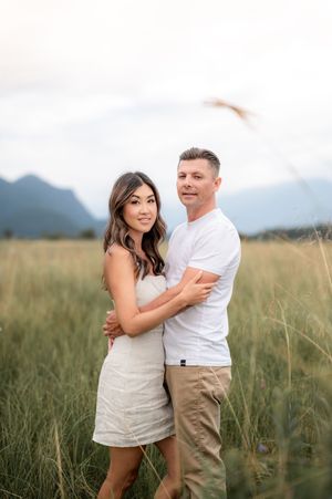 A romantic couple embraces in a grassy field with mountains visible in the background at sunset.