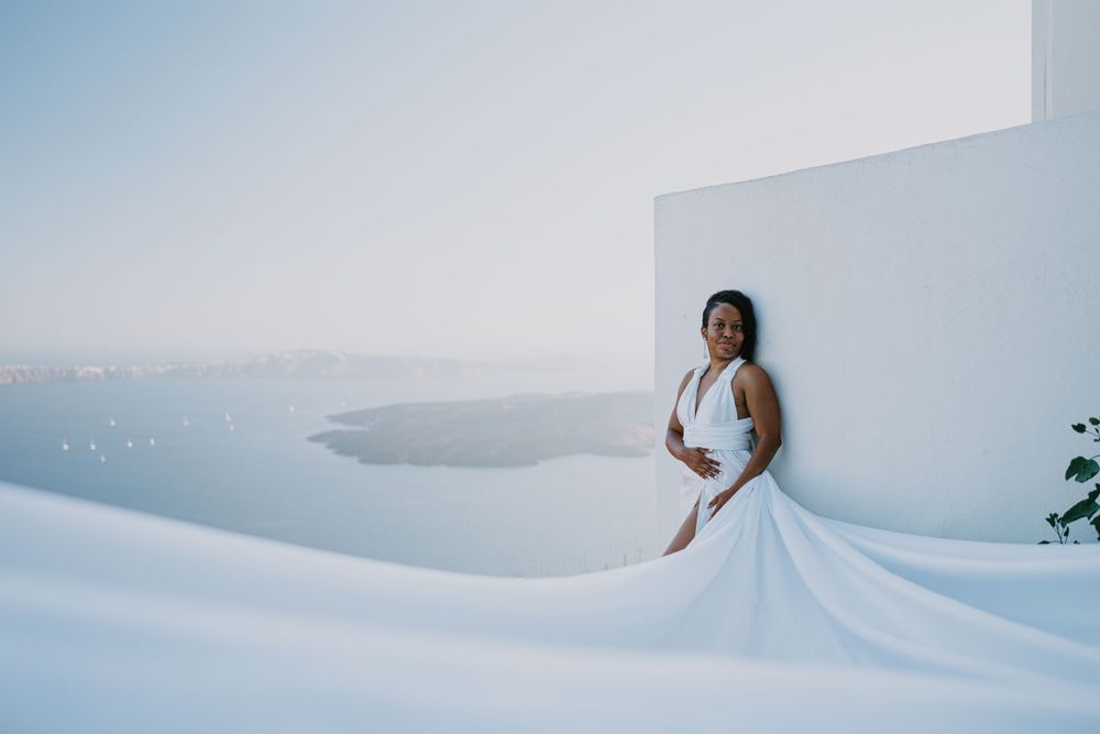 An elegant white dress flows against stark white walls with the blue Aegean Sea in the background.