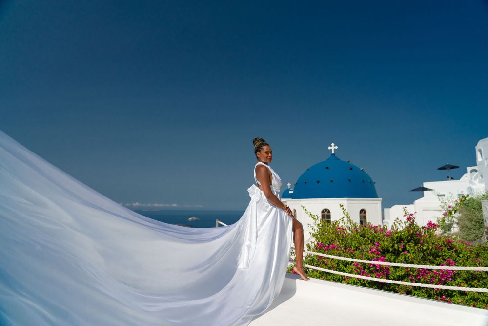 A dramatic white wedding dress trails in the wind against classic Santorini architecture.