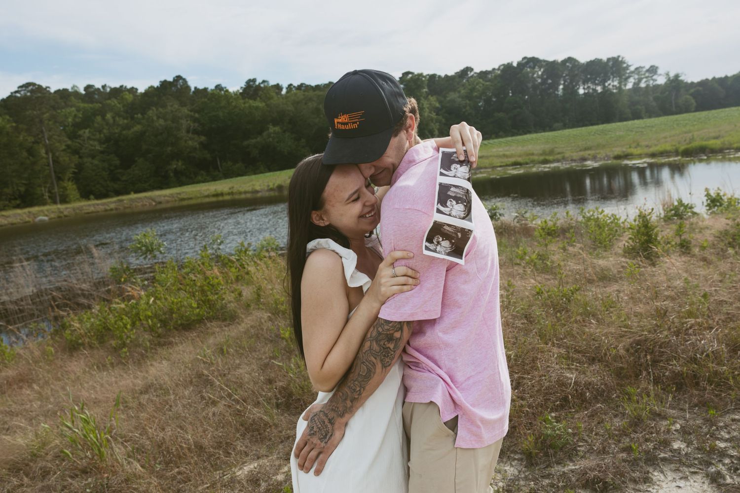 A couple embraces by a serene lake on a sunny day, surrounded by natural landscape.