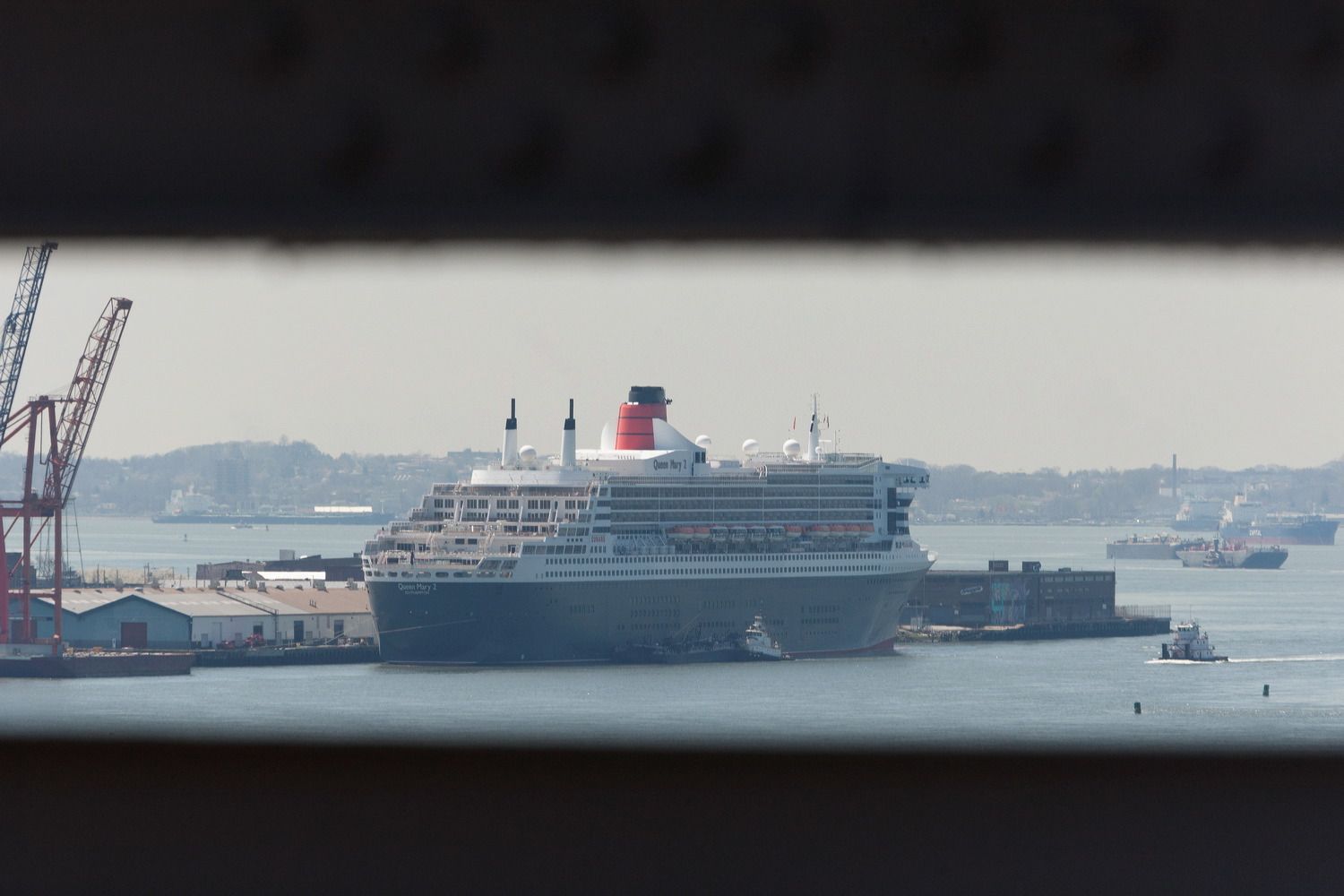 Queen Mary 2 cruise ship docked at port with cargo cranes visible in background.