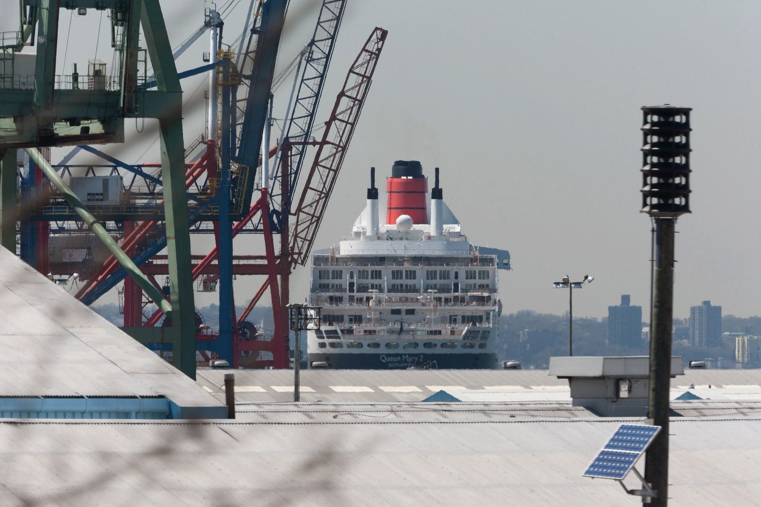 Queen Mary 2 cruise ship funnel visible among industrial port cranes and equipment.