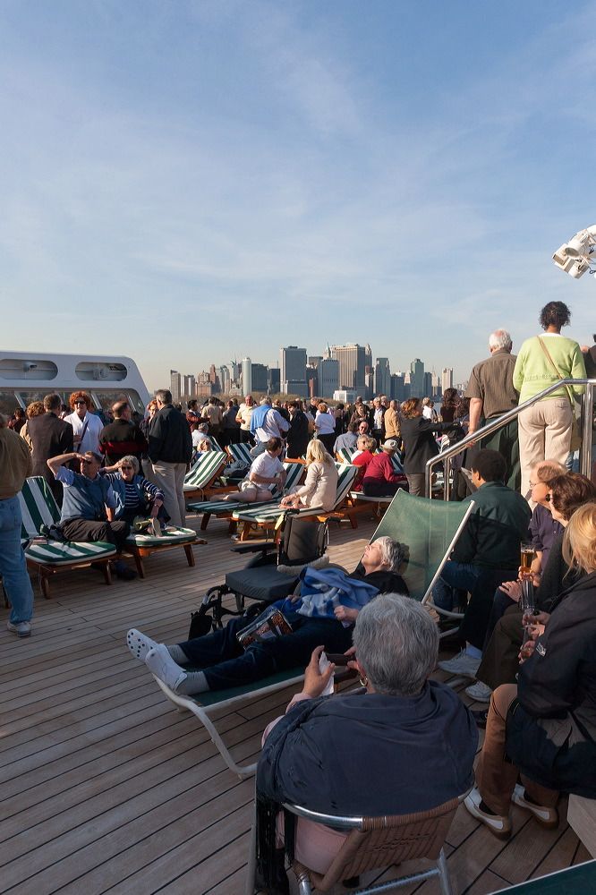 Cruise ship passengers lounging on deck chairs with Manhattan skyline visible in background.