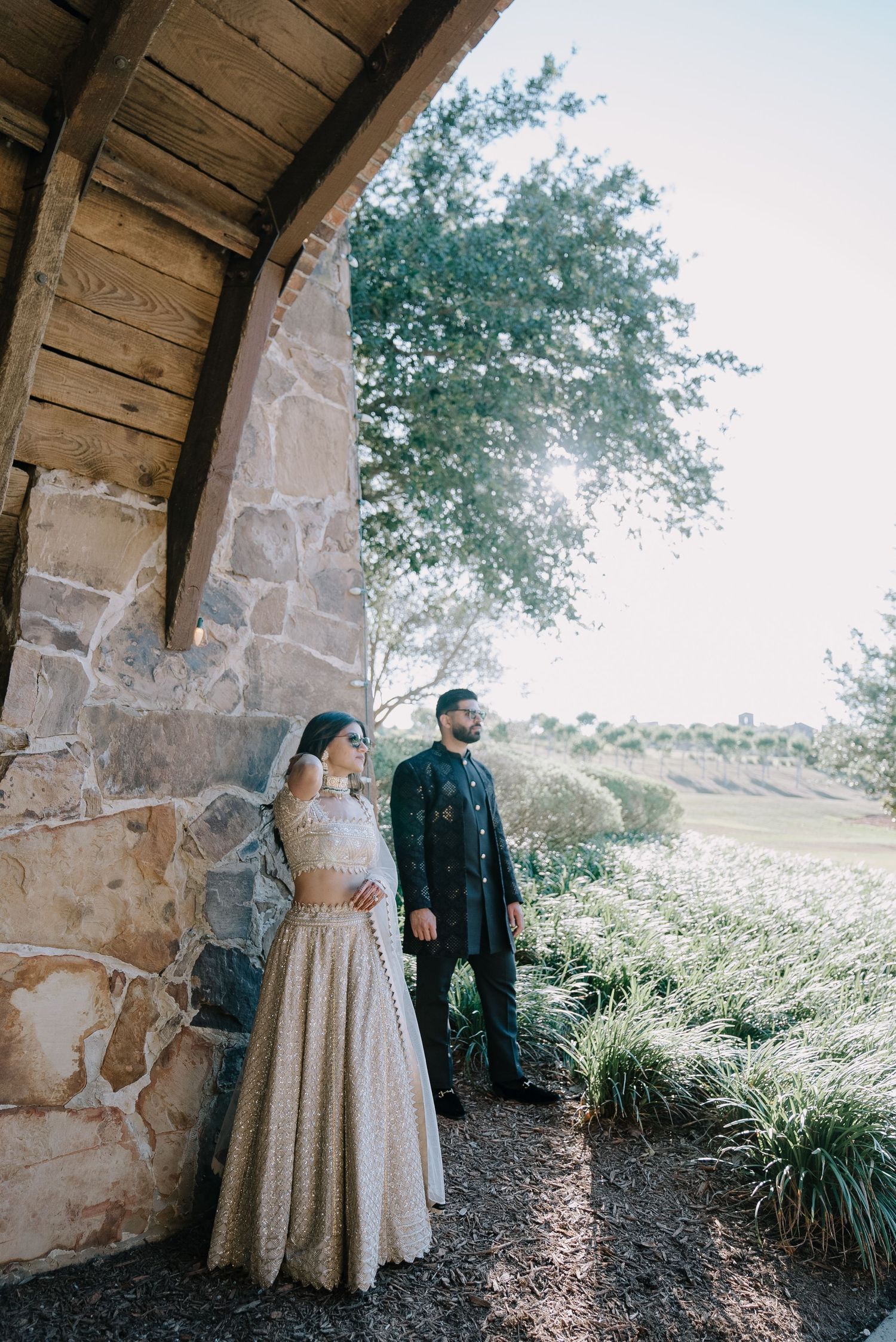 Couple poses under stone archway in elegant wedding attire amid scenic outdoor setting.