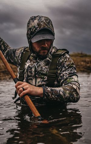 Hunter in camouflage gear wading through dark water with wooden paddle.
