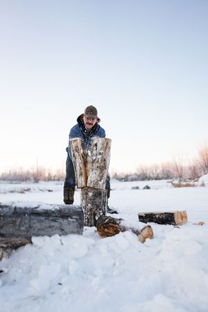 Person chopping wood in snowy winter landscape with scattered logs.