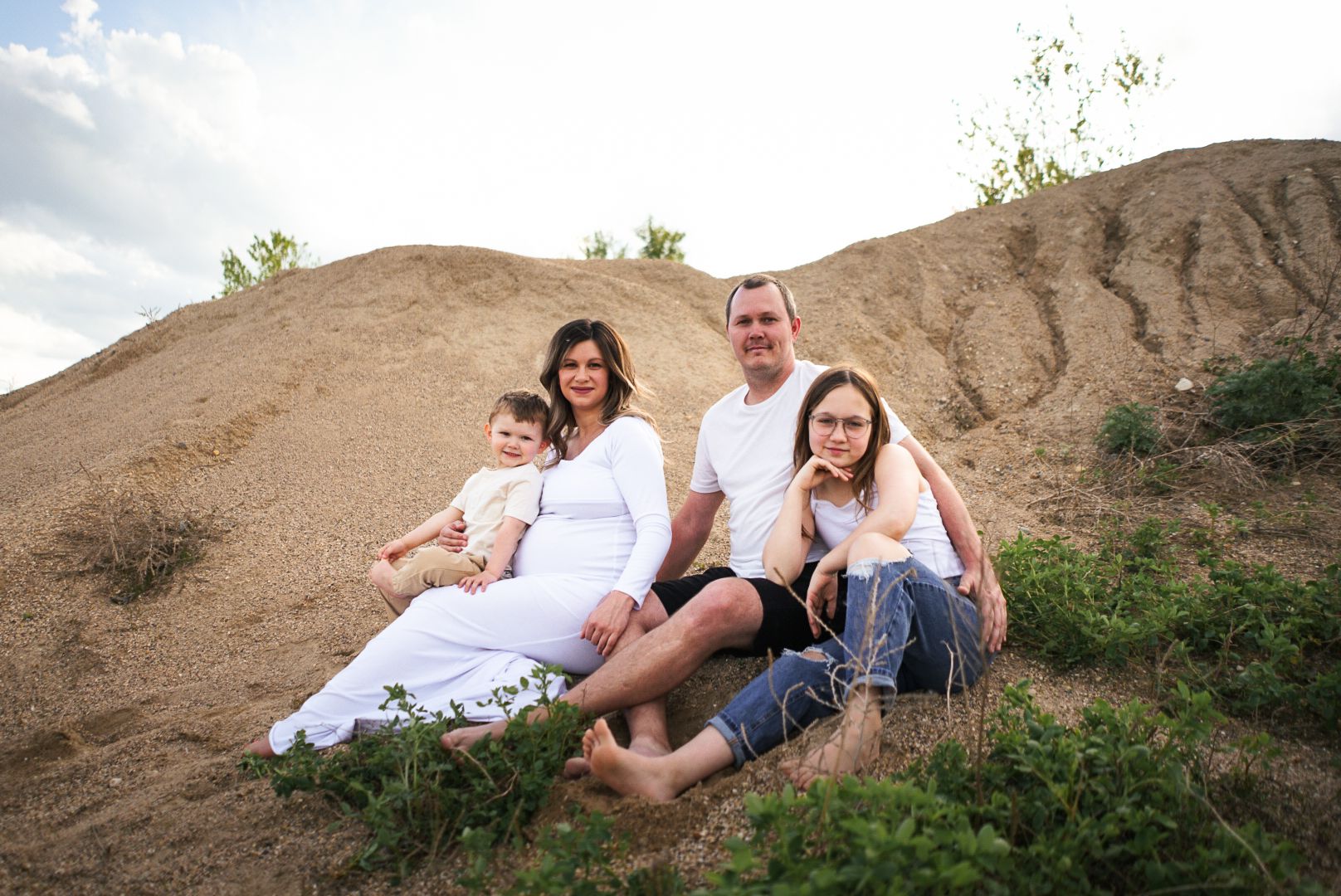 A family wearing white sits together on a sandy hill during an outdoor photo session.