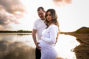 A couple embraces by a serene lake at sunset during a maternity photo session.