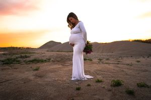 A person in a white dress stands against a desert sunset during a maternity photoshoot.