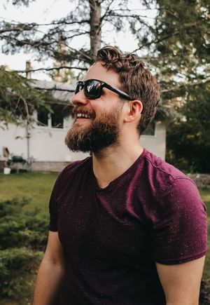 Man in sunglasses and purple shirt smiling while looking to the side outdoors.