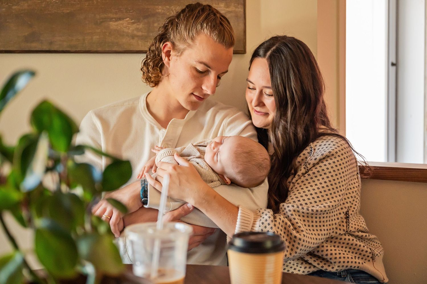 Two people share a tender moment while feeding a baby in a cozy home setting with houseplants and coffee nearby.