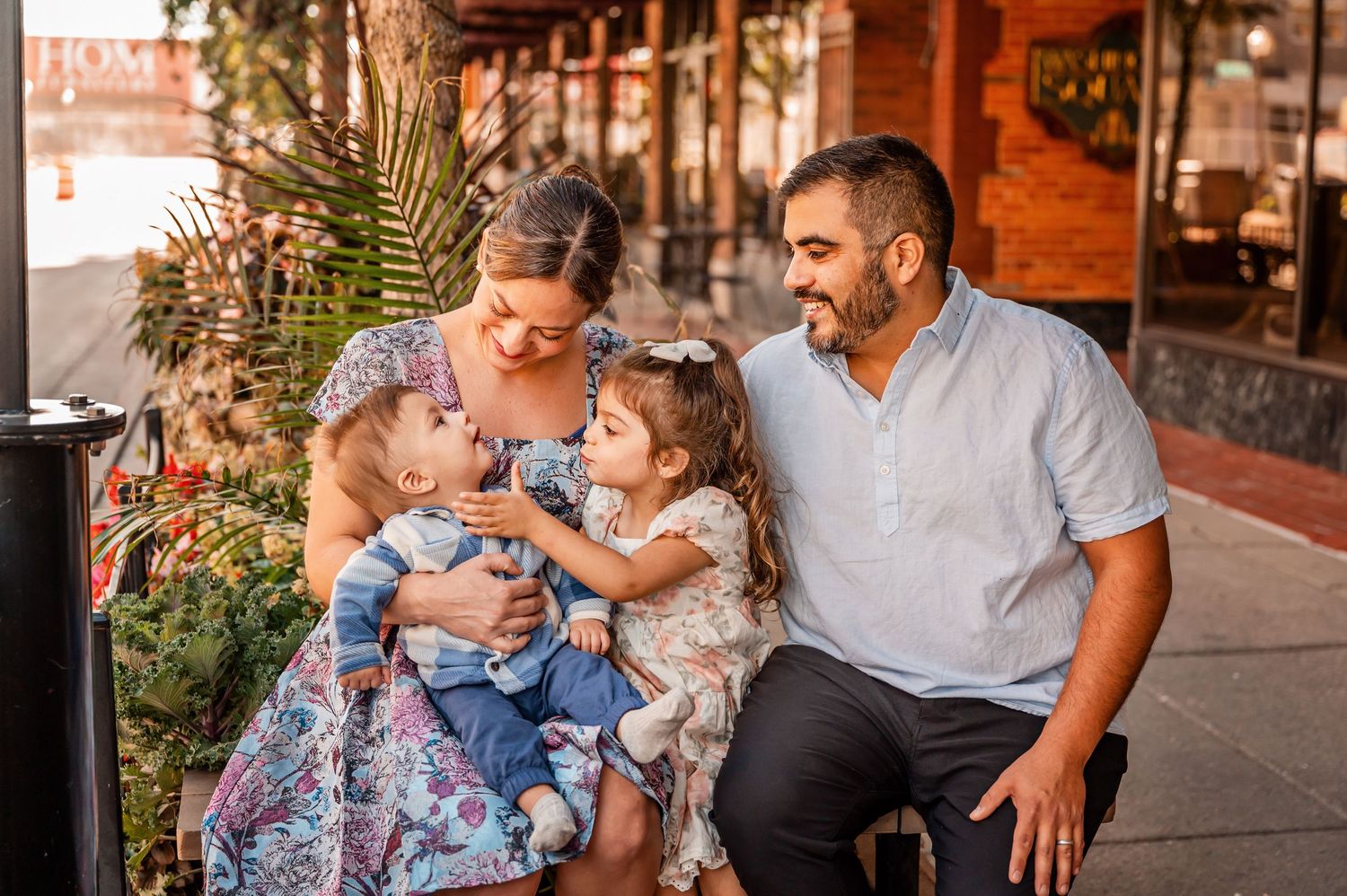 A family sits together on a city sidewalk near decorative plants, sharing a sweet moment on a sunny afternoon.