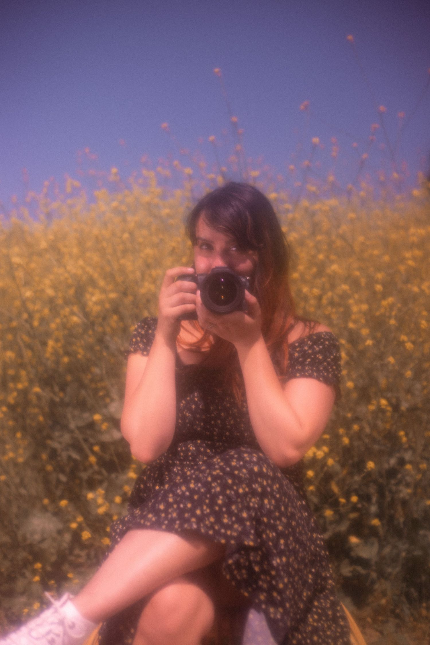 Kelsey Nevell holds a camera to her face with yellow flowers in the background.