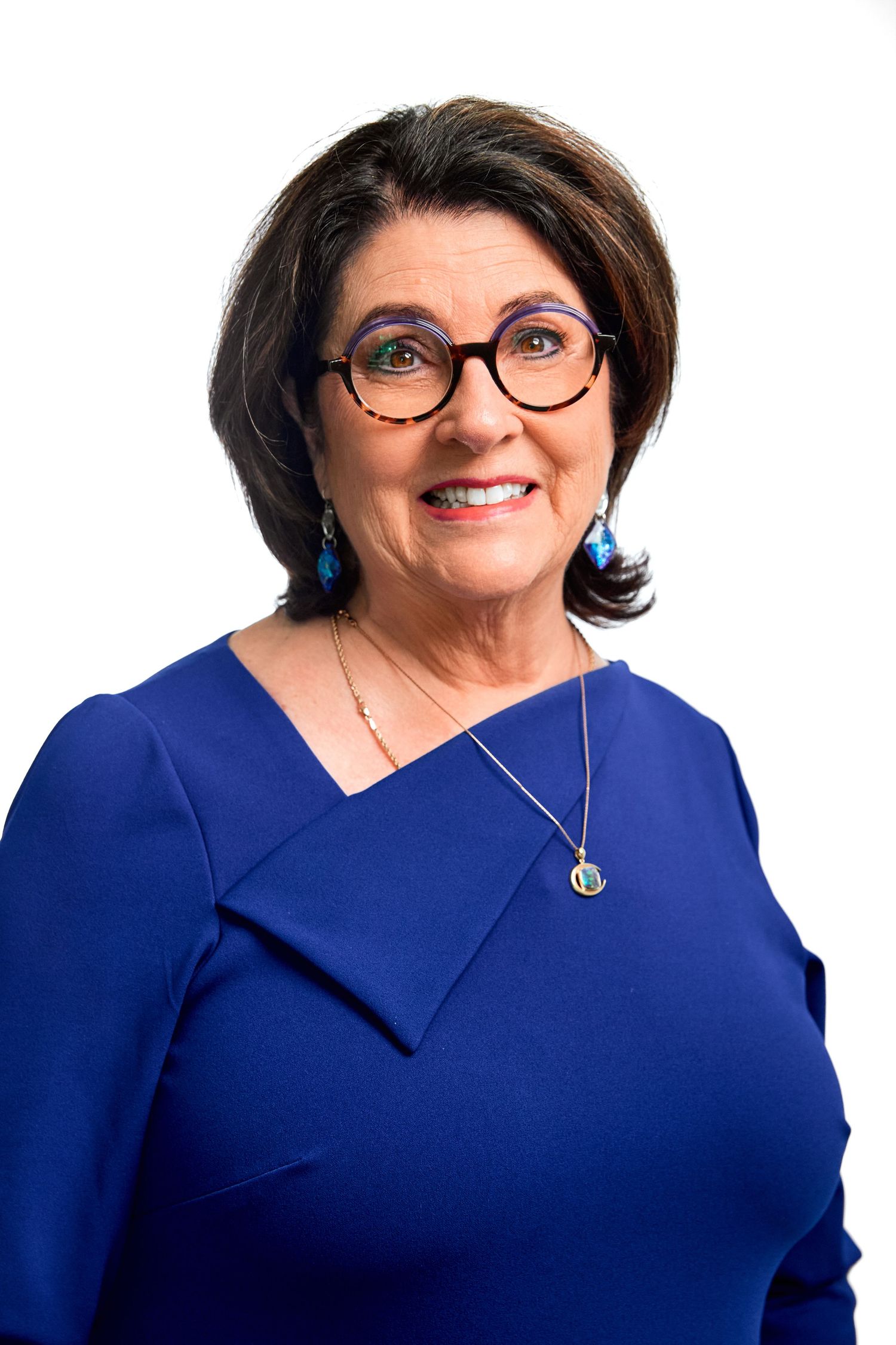 Smiling older woman with brown hair and glasses in blue dress with necklace on white background.