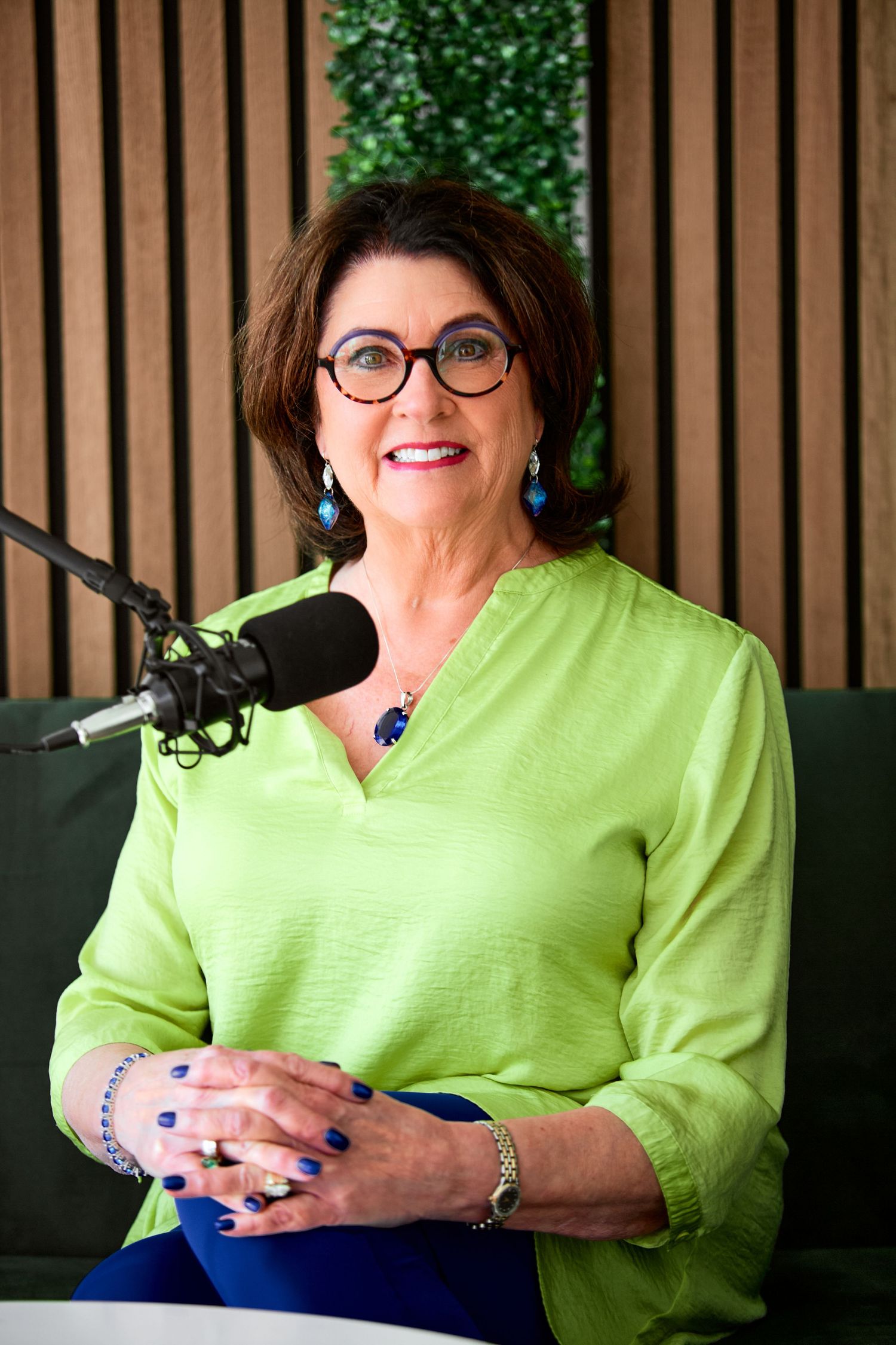Smiling older woman with glasses in green blouse sitting at podcast microphone in modern studio.