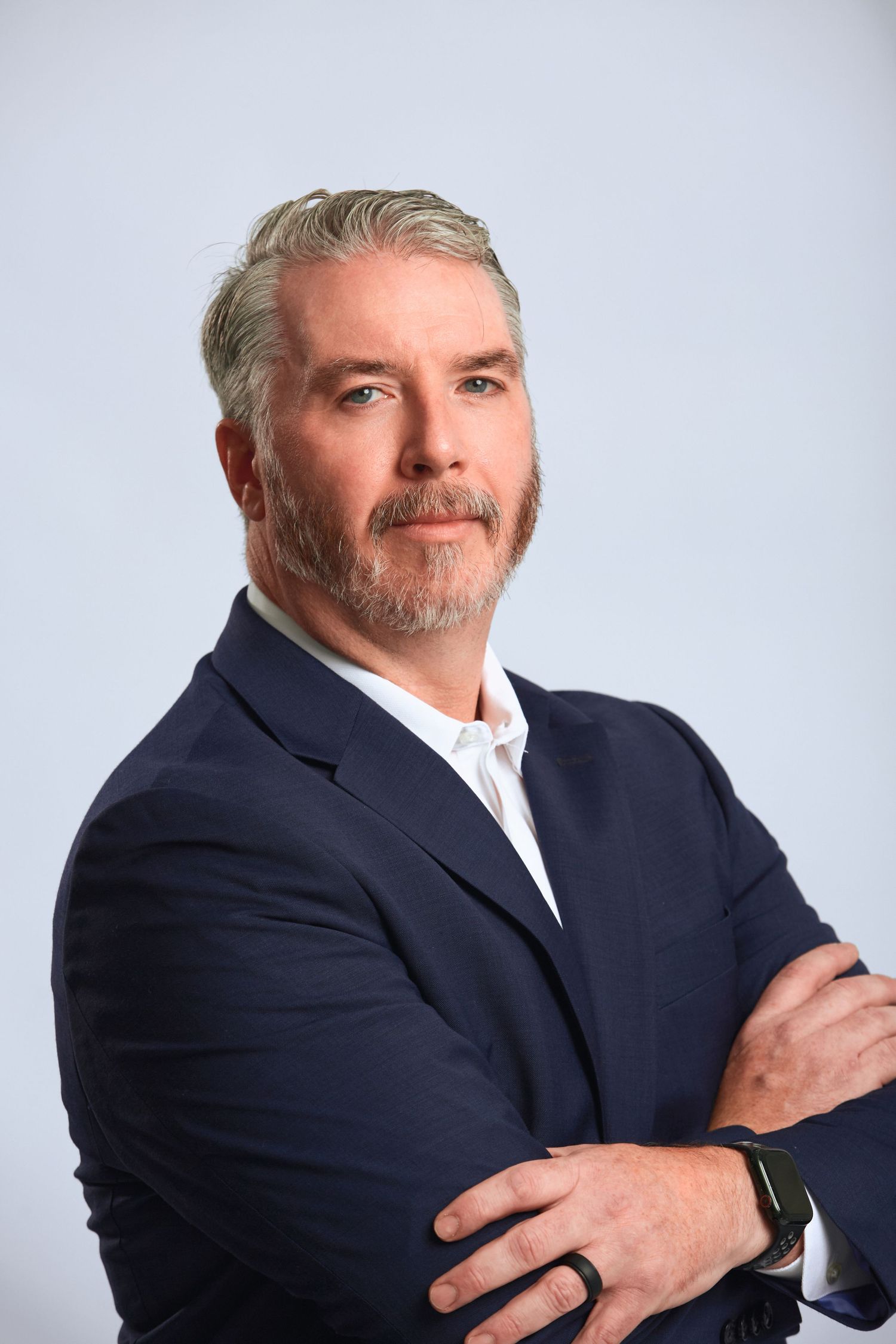 Confident man with gray hair and beard in navy blazer with arms crossed against blue background.