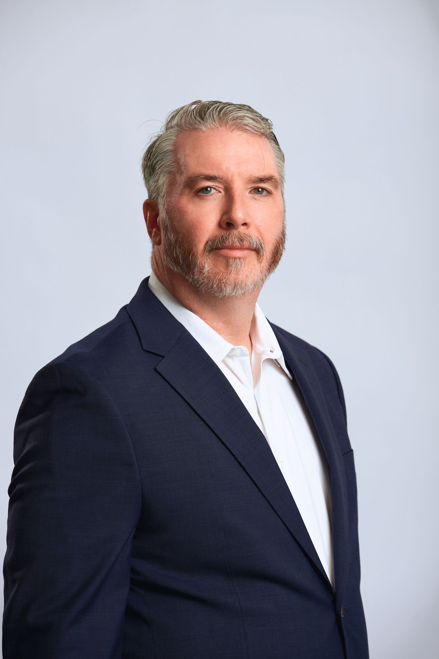 Middle-aged man with gray hair and beard in navy blazer posing against light blue background.