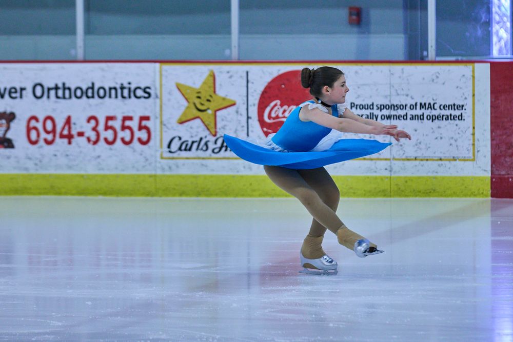 A figure skater performs a graceful pose in a blue dress on an ice rink with advertising boards visible in the background.