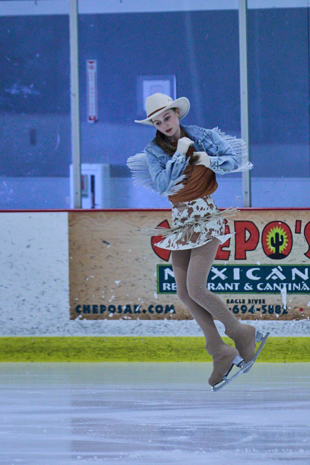 A figure skater performs an artistic spin on ice while wearing a denim jacket and patterned skirt in front of rink advertisements.