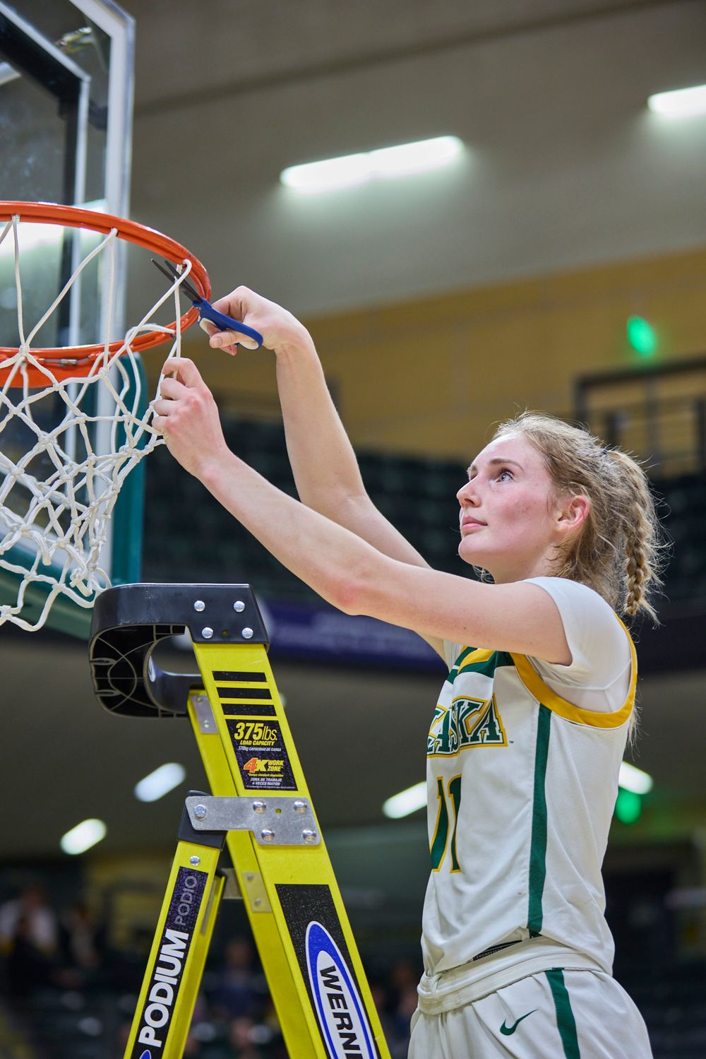 Basketball player cuts down championship net from atop yellow ladder.