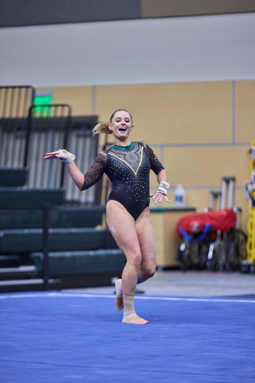 Gymnast in black sparkly leotard performs dance movements on blue floor mat.