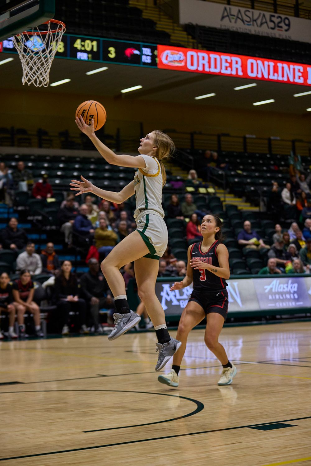 Basketball player leaps for a layup during a competitive indoor game while opponent defends.