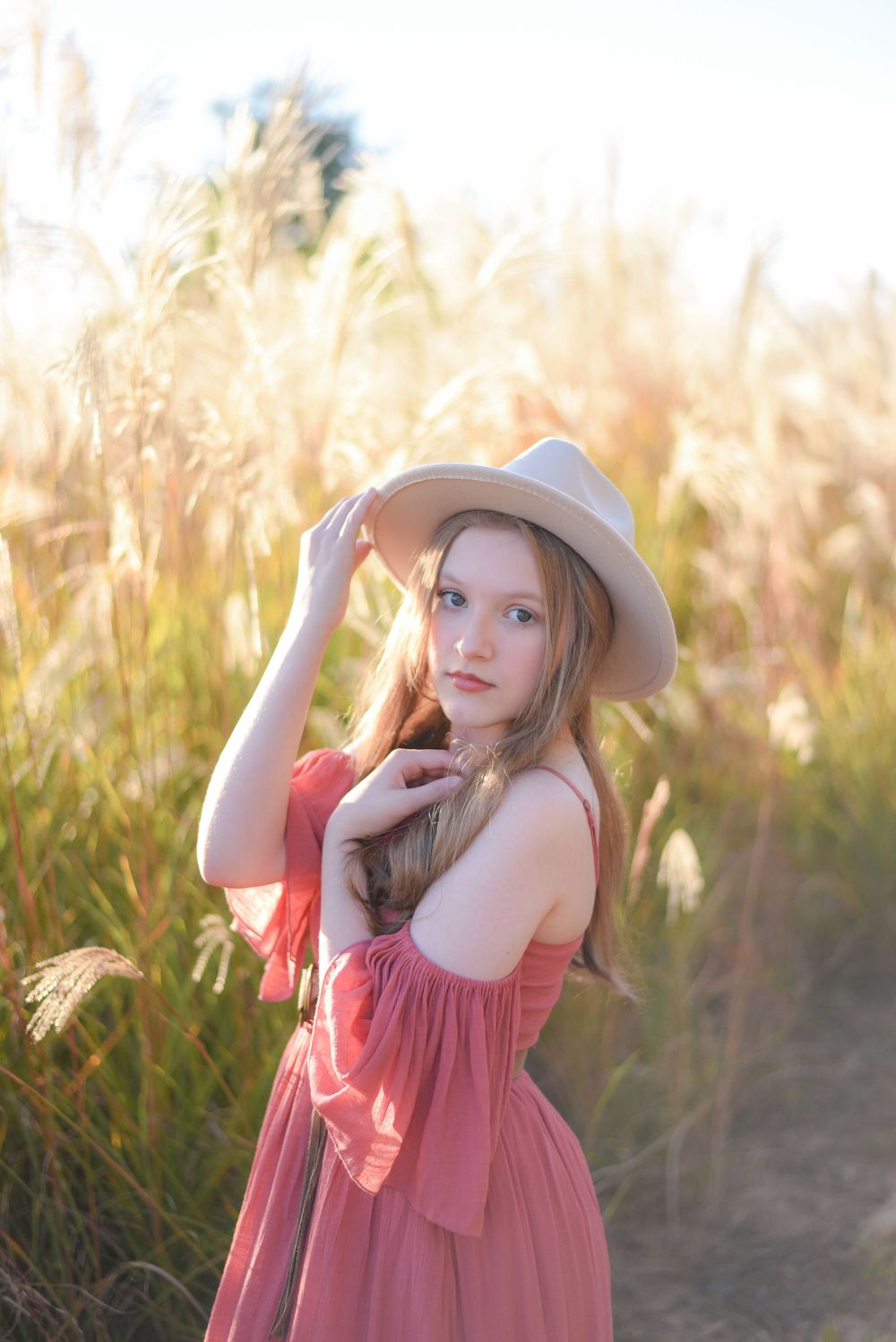 Whimsical poses in pink dress and hat against sunlit field of tall grass.