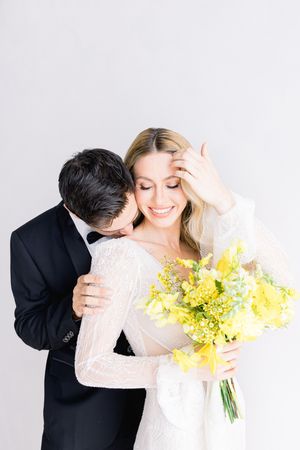 Tender wedding moment with couple embracing as bride holds vibrant yellow spring bouquet against white background.