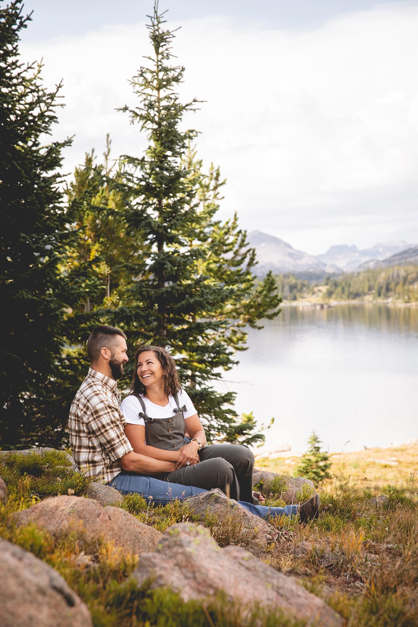 husband and wife sit in the shade from pine trees near a mountain lake