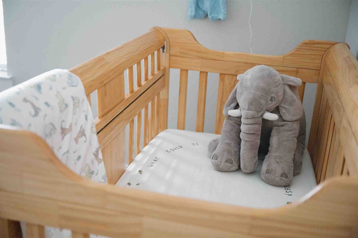 A wooden baby crib with white bedding and a plush gray elephant toy resting against the side rail.