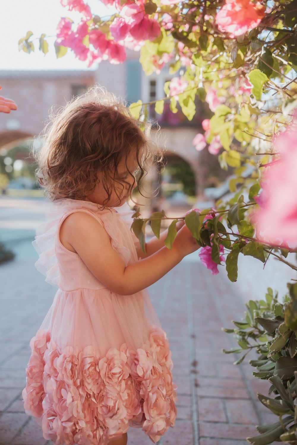 A child in a pink dress reaching up to touch pink bougainvillea flowers.
