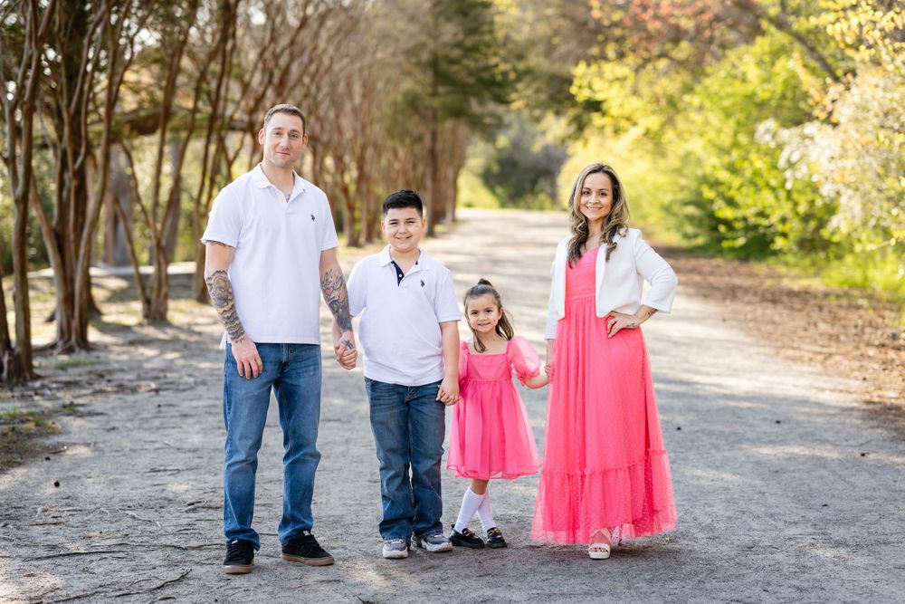 A family of four poses on a tree-lined path in coordinated pink and white outfits.