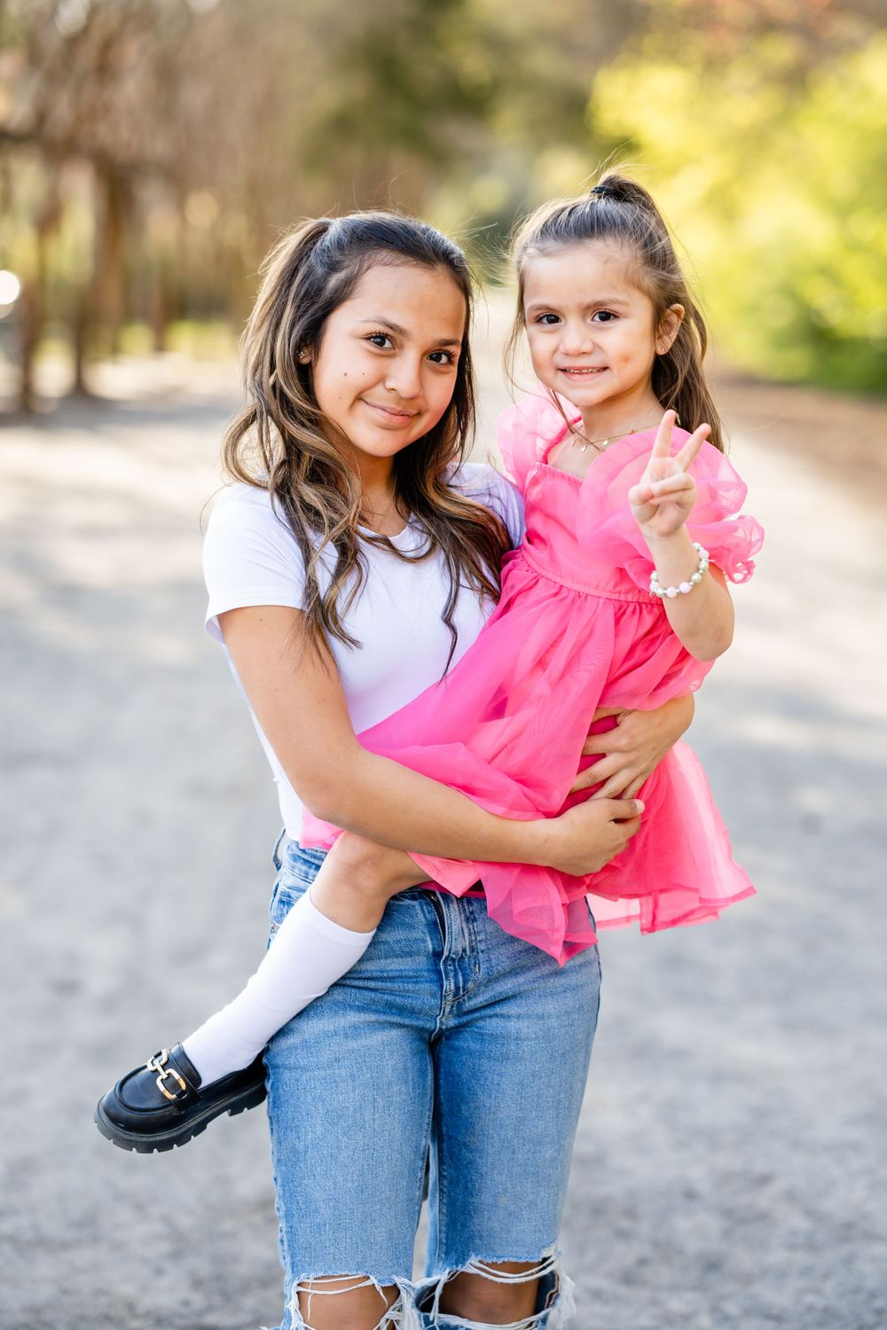 Siblings sharing a tender moment on a wooded path as the older sister holds her little sister wearing a pink dress.