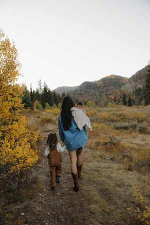 Mother carries baby while walking with young daughter through a scenic autumn mountain landscape.