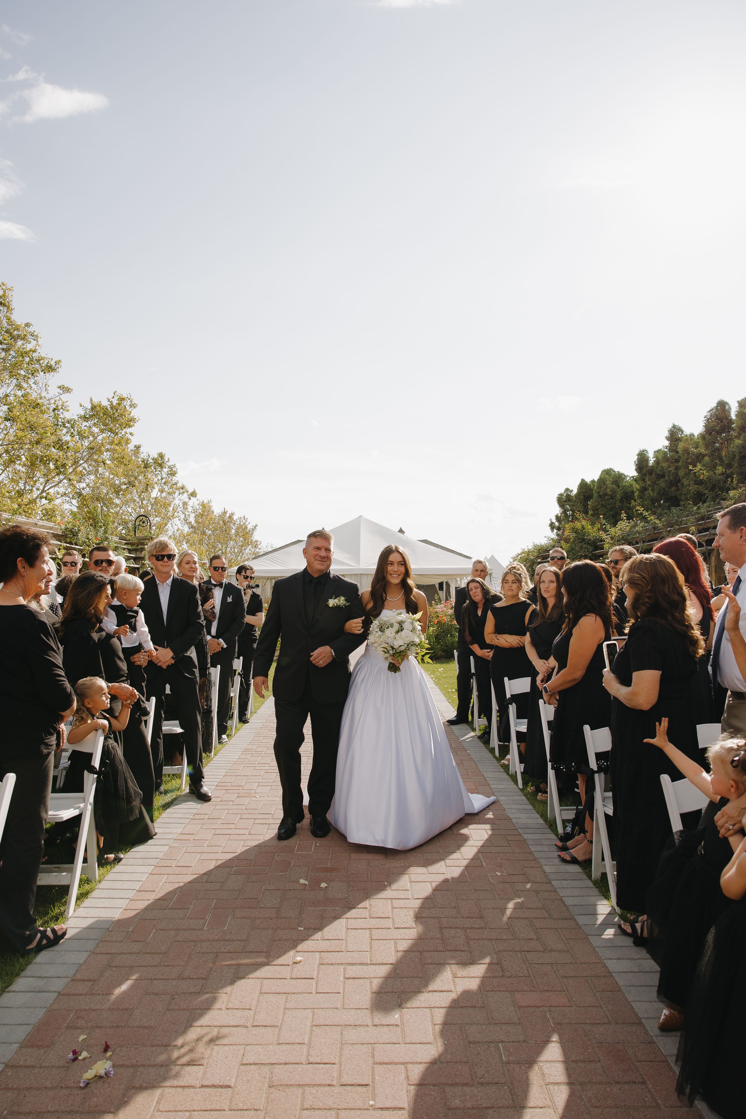 Bride walks down the outdoor aisle with her escort as guests in black attire look on at the ceremony.