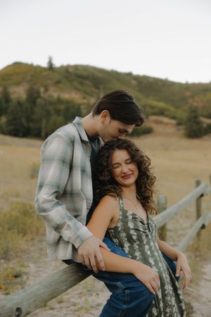 Young couple poses together on a rustic fence in a golden hillside meadow at dusk.