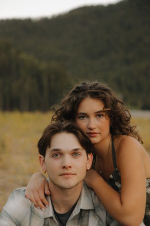 Young couple posing outdoors in a mountain meadow, woman with curly hair embracing man from behind.