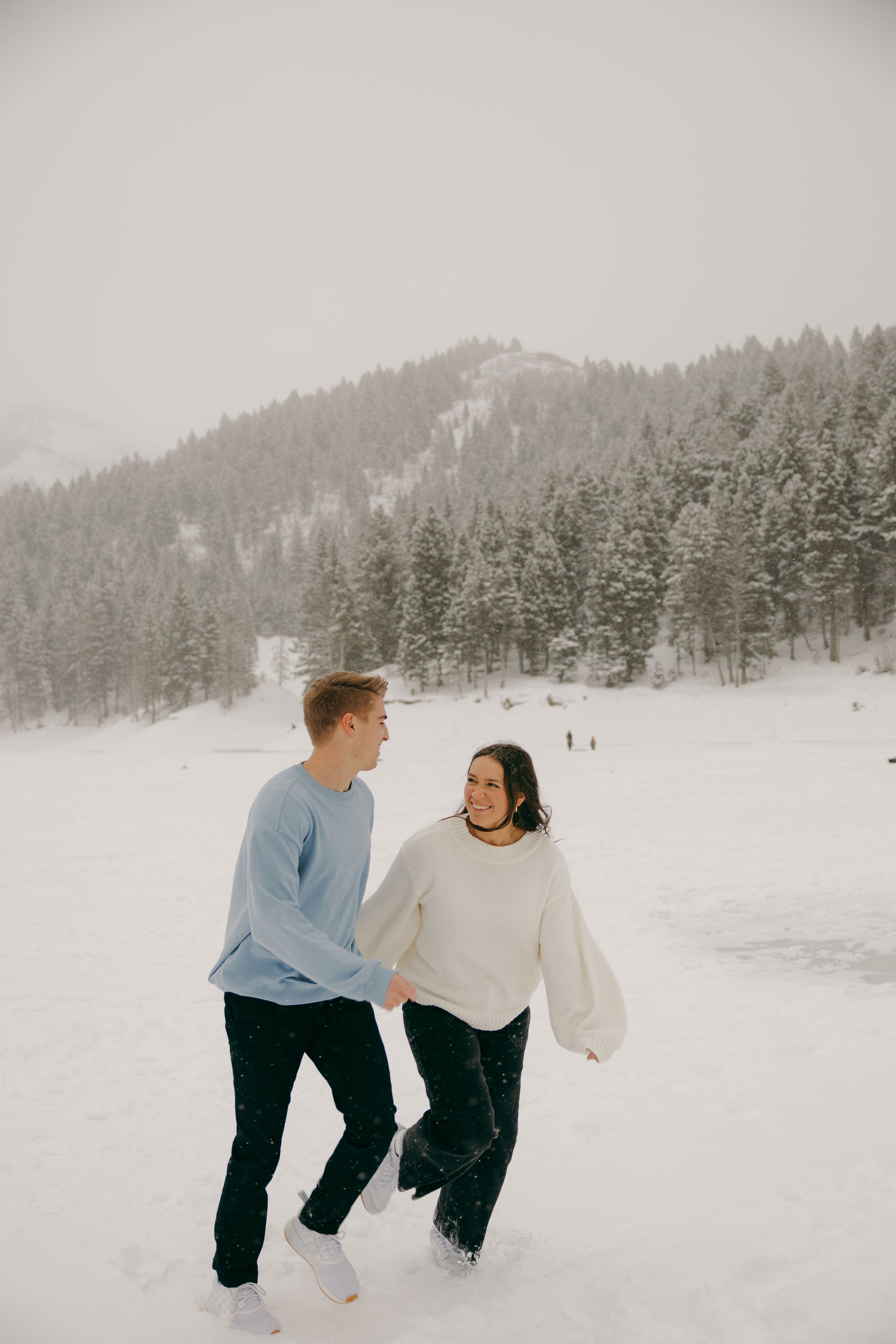 Couple holding hands and walking through a snowy winter landscape with snow-covered pine trees in the background.