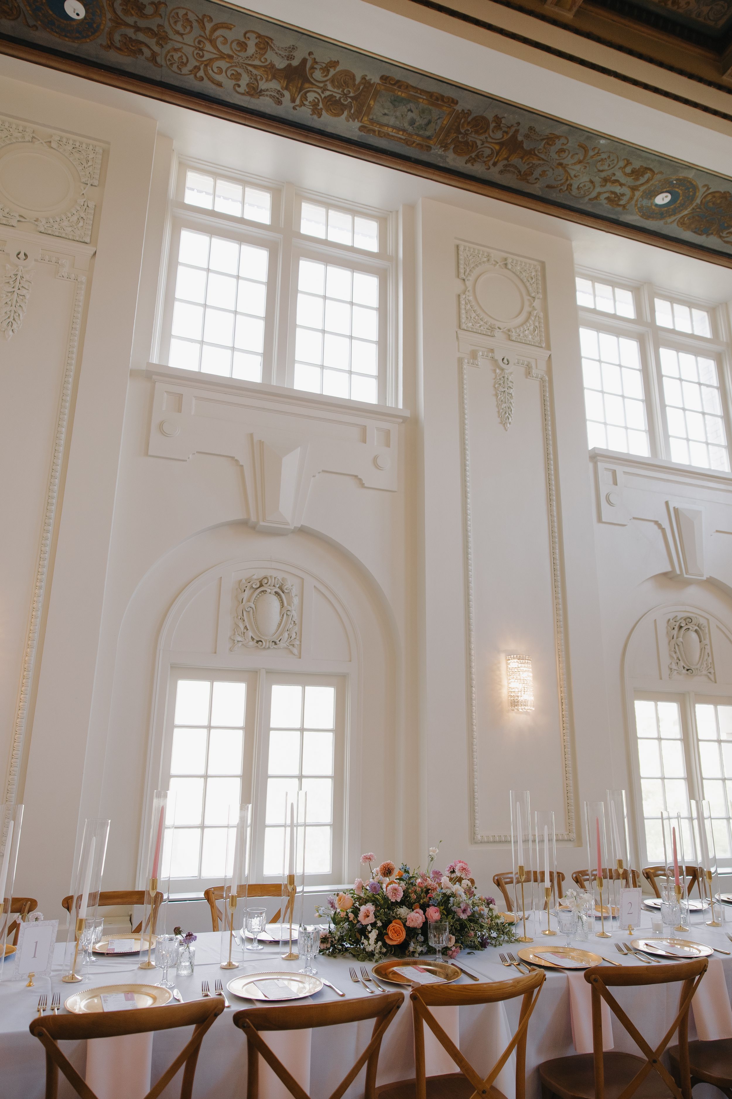 Elegant white wedding reception hall with ornate moldings, tall windows, and a floral centerpiece table.