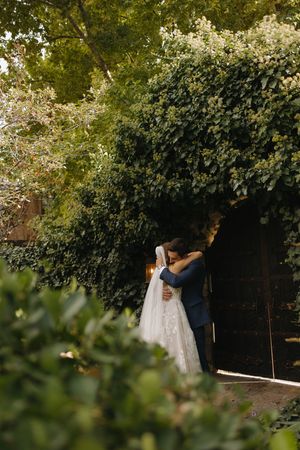 Bride and groom share a tender embrace under a lush ivy-covered archway at a romantic garden venue.