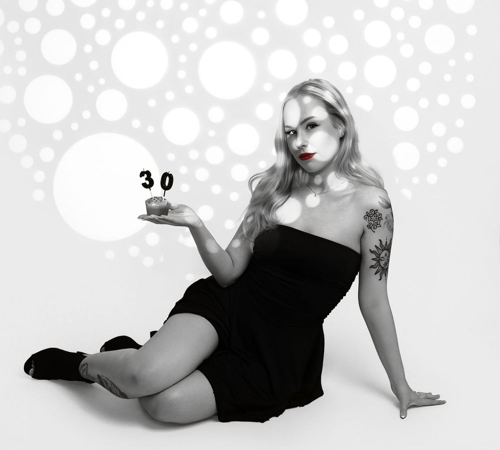 Black and white portrait of someone in a short black dress posing with a number 30 against a polka dot background.
