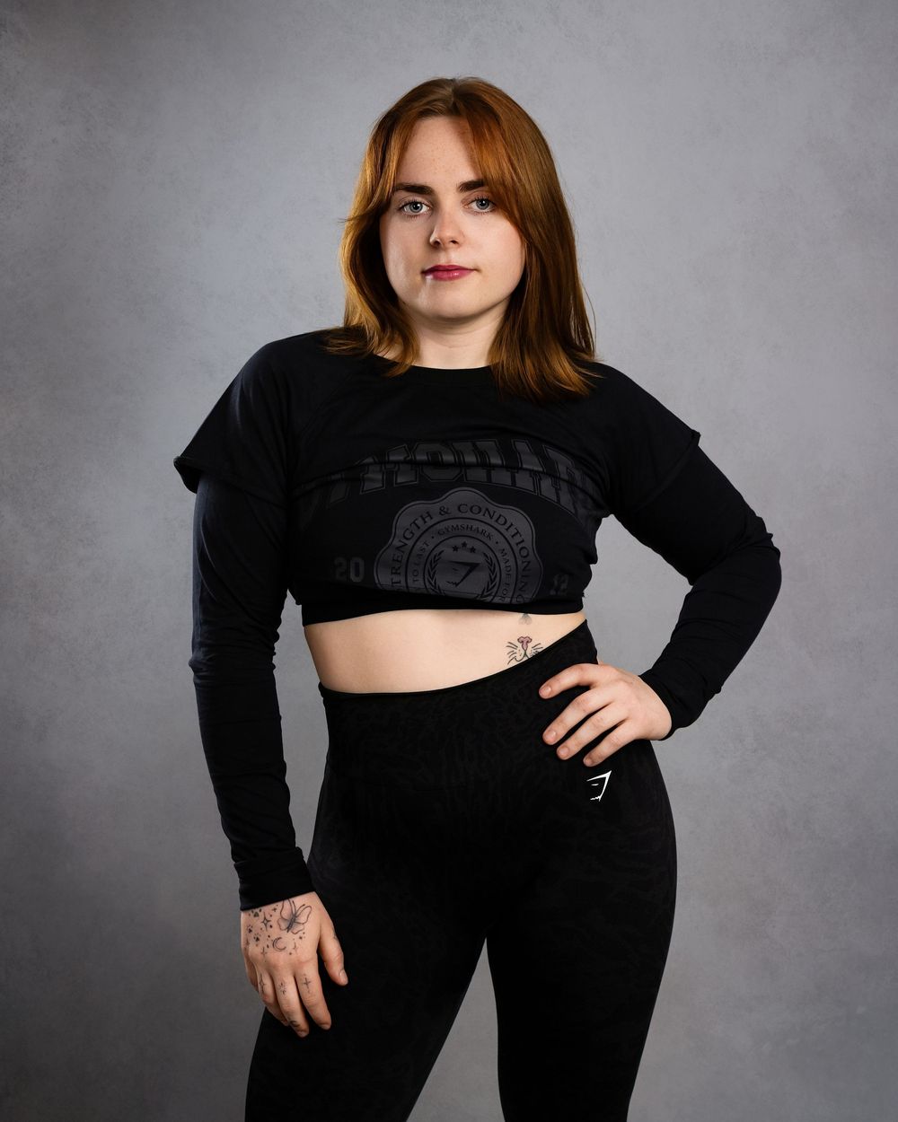 Woman in black crop top and leggings poses confidently with hand on hip against gray background.