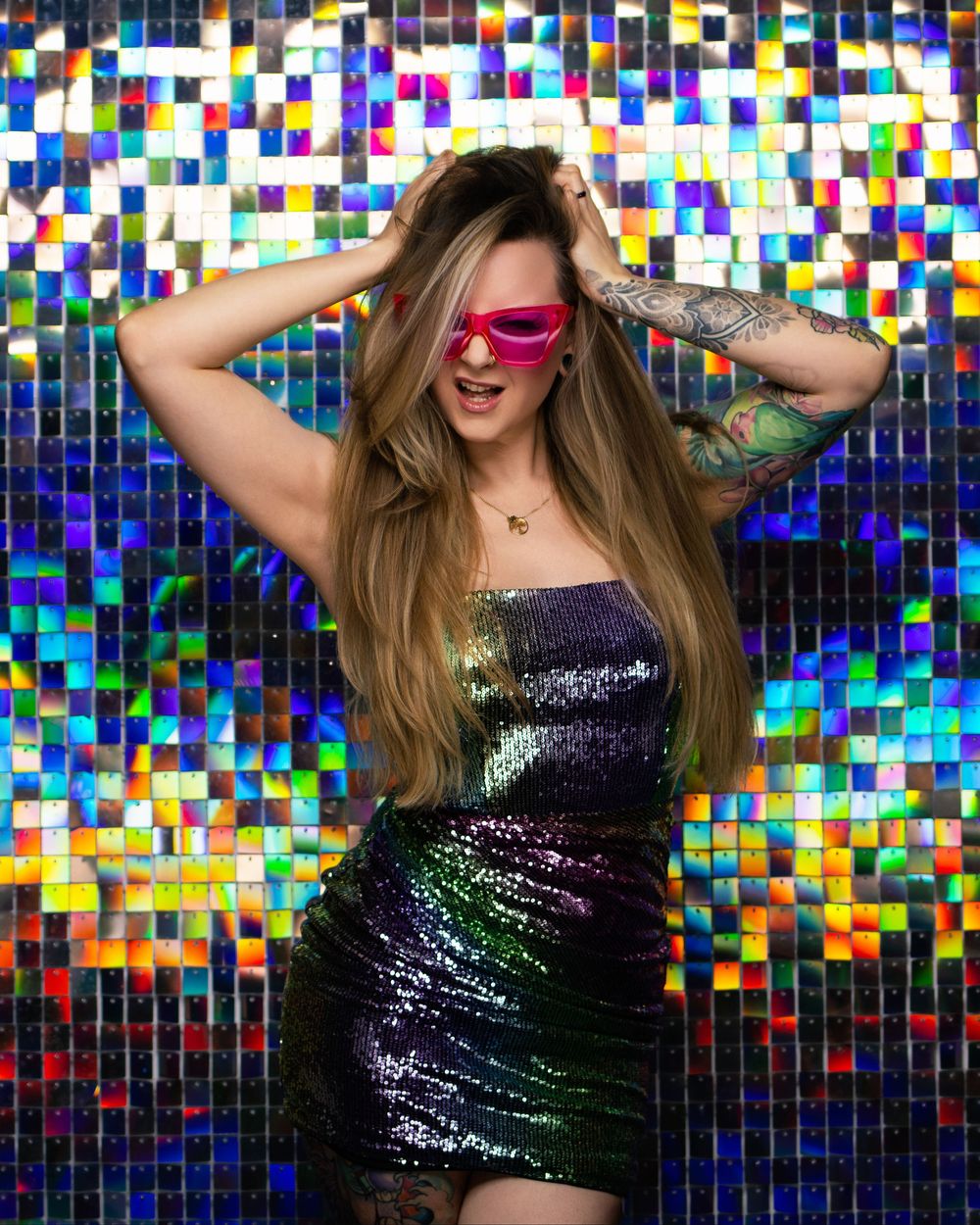 A person in a sparkly black dress poses playfully against a colorful mosaic wall with blue and rainbow tiles.