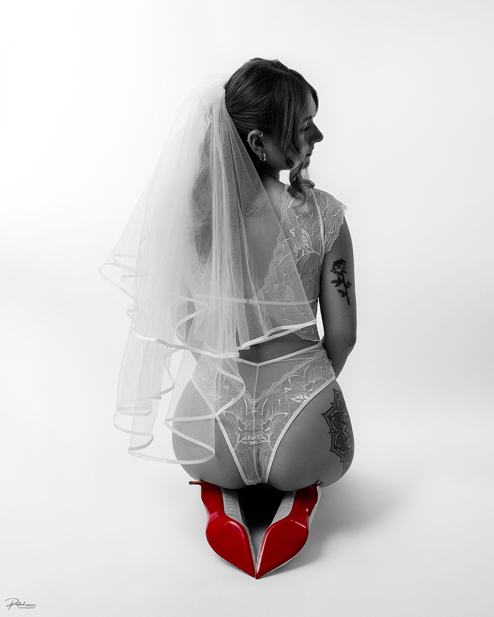 Woman in bridal lingerie and veil kneeling in red heels, black and white photo with selective color.