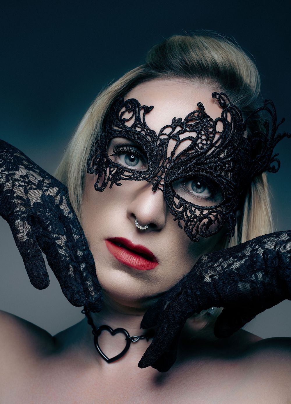 Blonde woman wearing black lace masquerade mask and gloves with red lips and heart necklace.