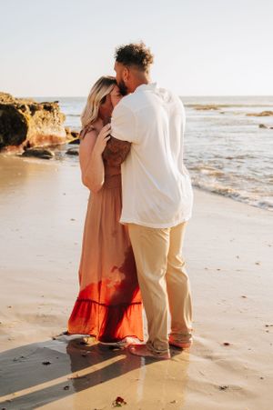 A romantic couple embraces on a beach at sunset with rocky cliffs in the background.