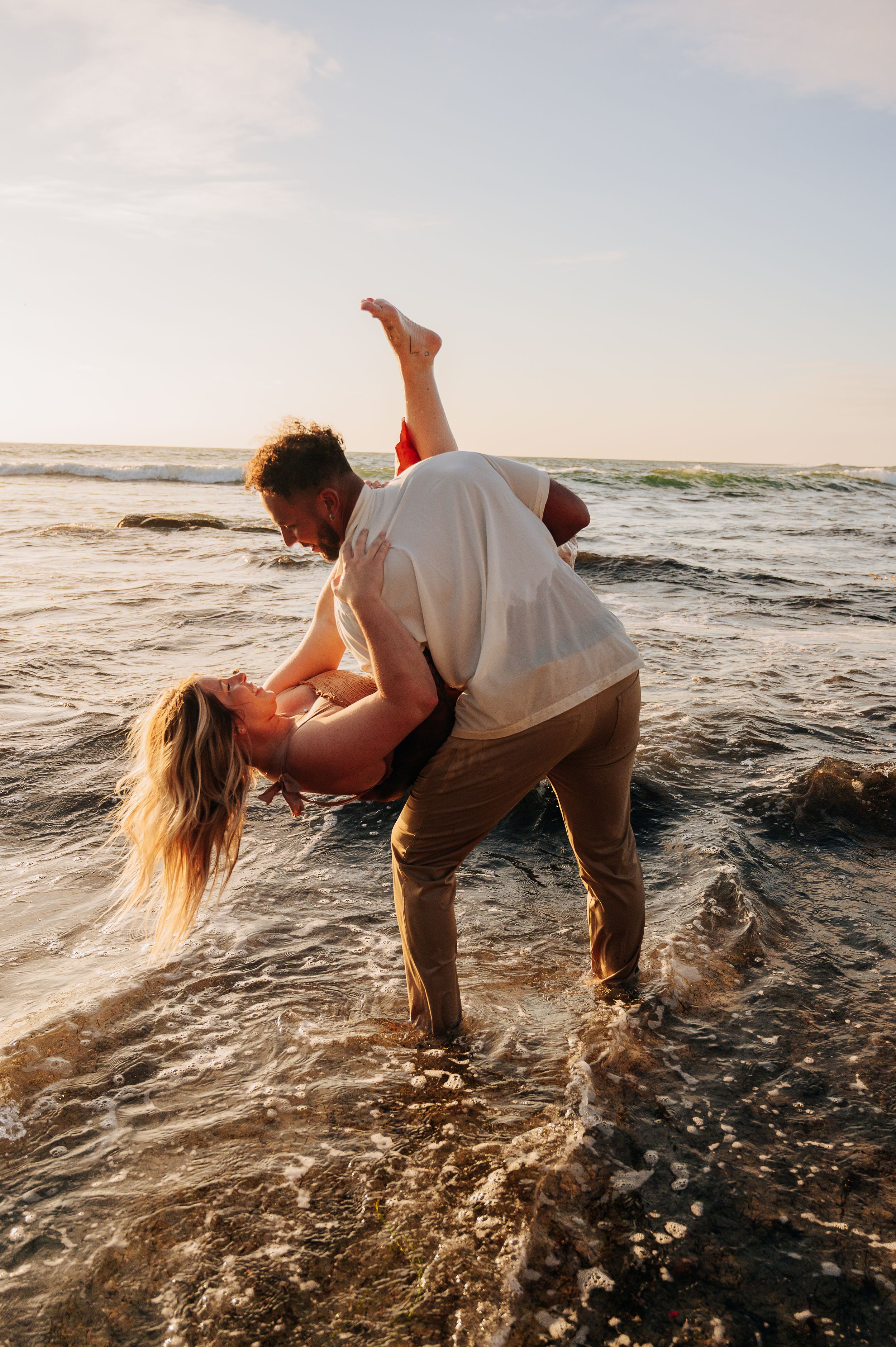 A playful couple dances in the ocean waves during sunset.